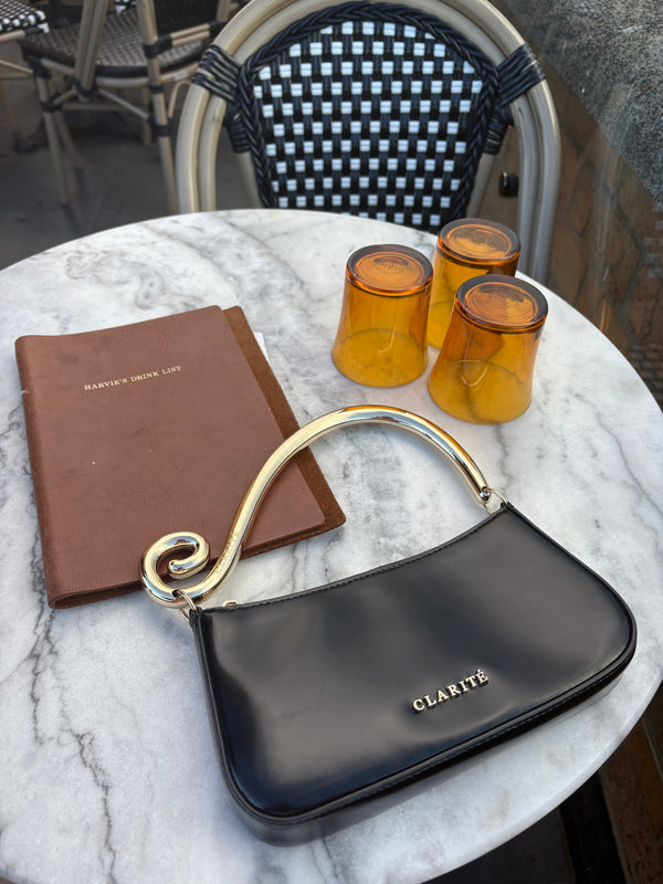 Black handbag with gold handle, brown wallet, and amber glasses on a marble surface.